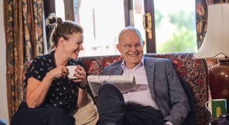 Woman and older man sitting on a sofa at home with a cup of tea and newspaper smiling