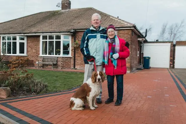 Older couple stand together outside their house with a dog