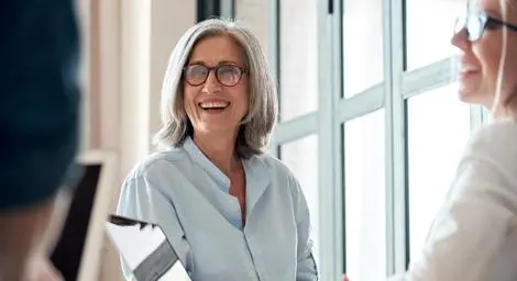 Older mature woman laughing and chatting with colleagues using laptops