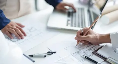 Two people are working over a set of blueprints, one is holding a pencil