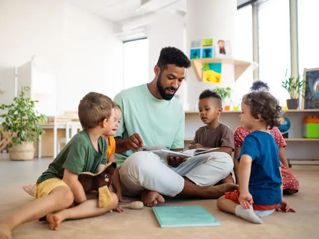 male adult sitting on the floor reading a book to a number of children.