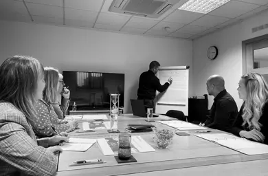 Black and white photograph of five people sitting in a meeting room with a tutor writing on a flipchart
