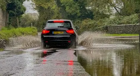 A black car drives through a flooded road, brown water splashes up from their tyers