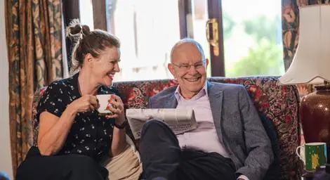 Woman and older man sitting on a sofa at home with a cup of tea and newspaper smiling