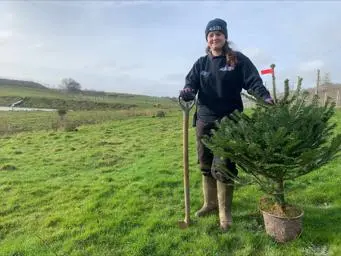 Katie Bowes next to pine tree sapling, holding a shovel and standing on a field