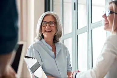 Older mature woman laughing and chatting with colleagues using laptops